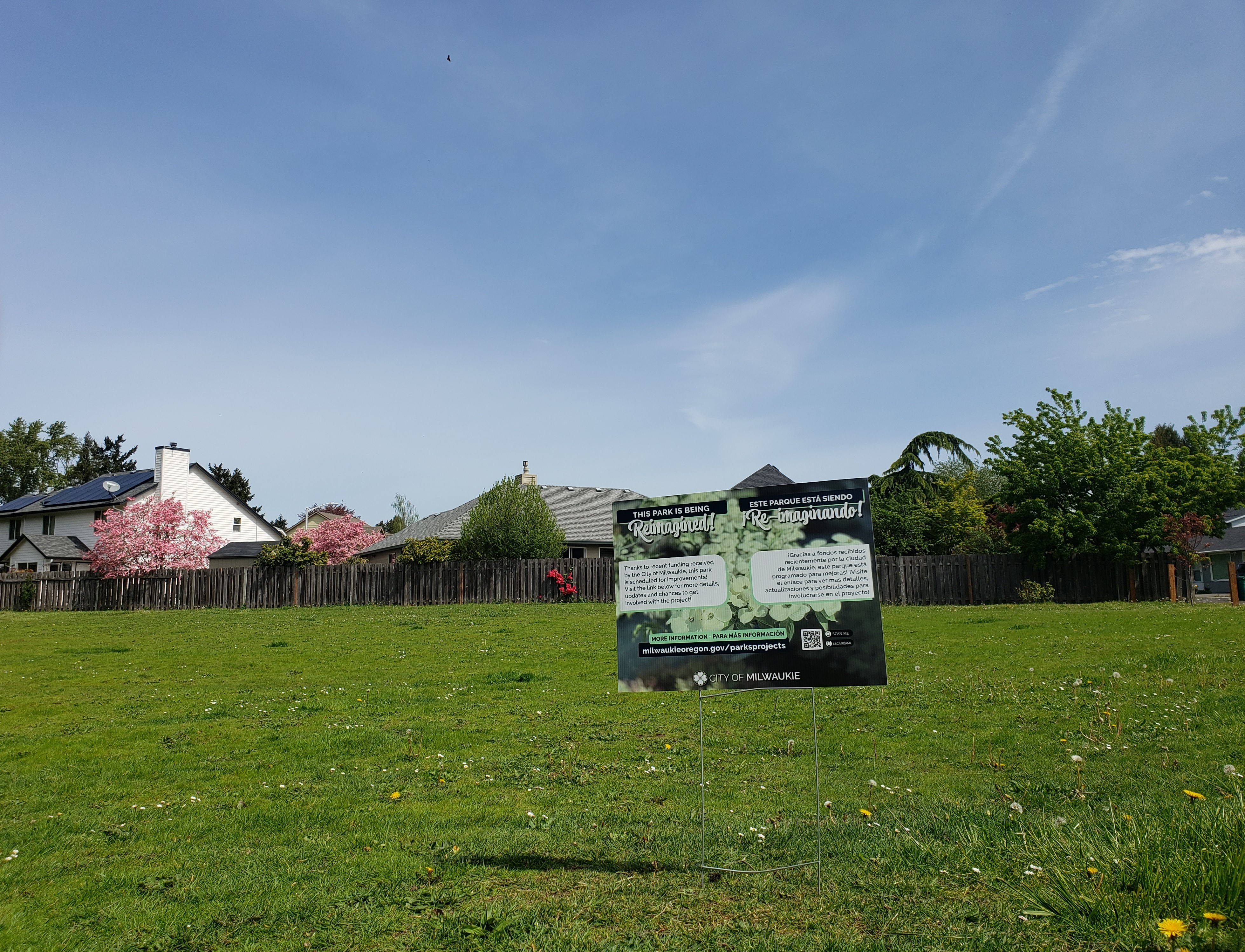  A yard sign explaining the park will be reimagined is on grass field under a blue sky with houses in the background.