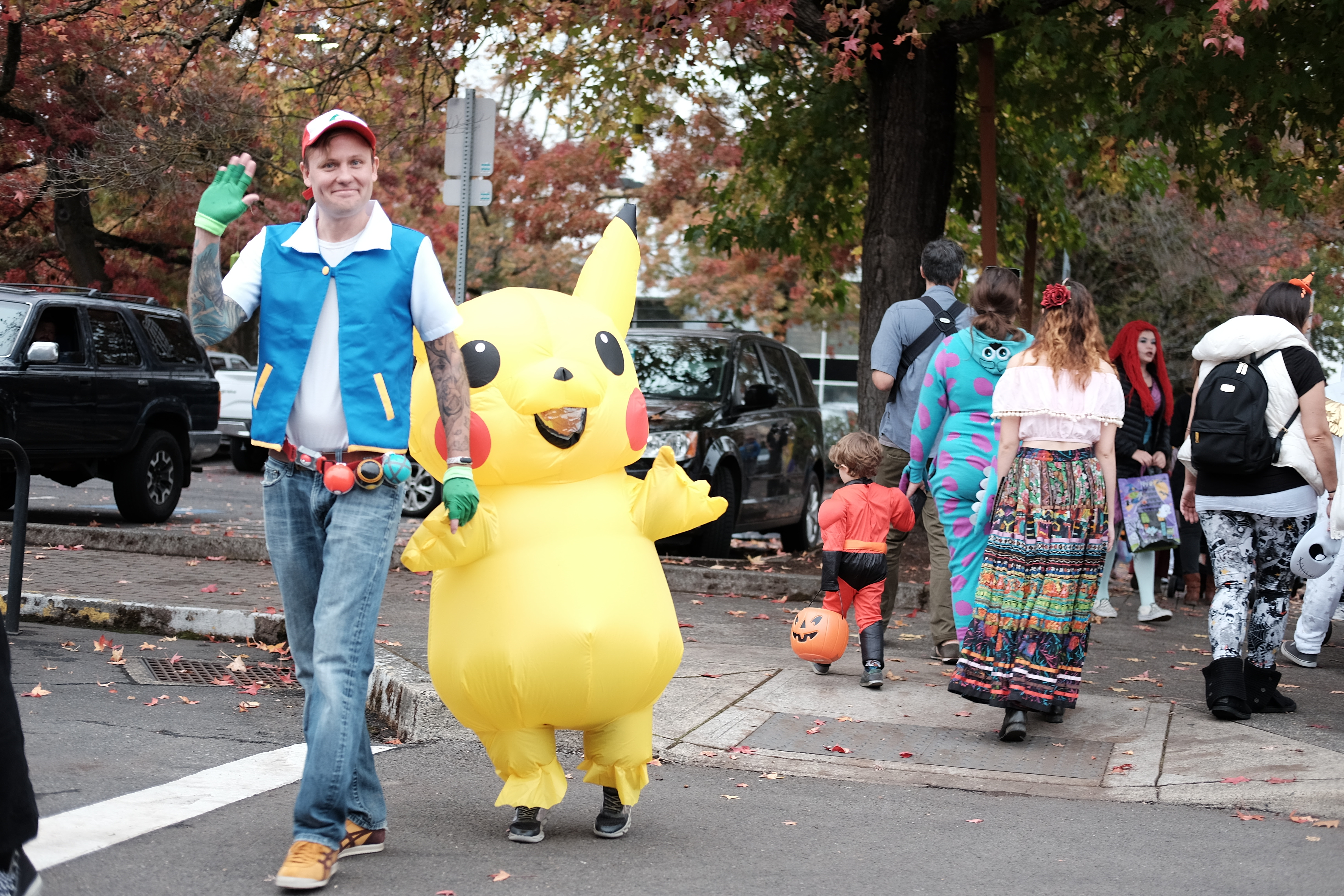 An adult dressed as Ash Ketchum leads a child dressed as Pikachu across the street. 